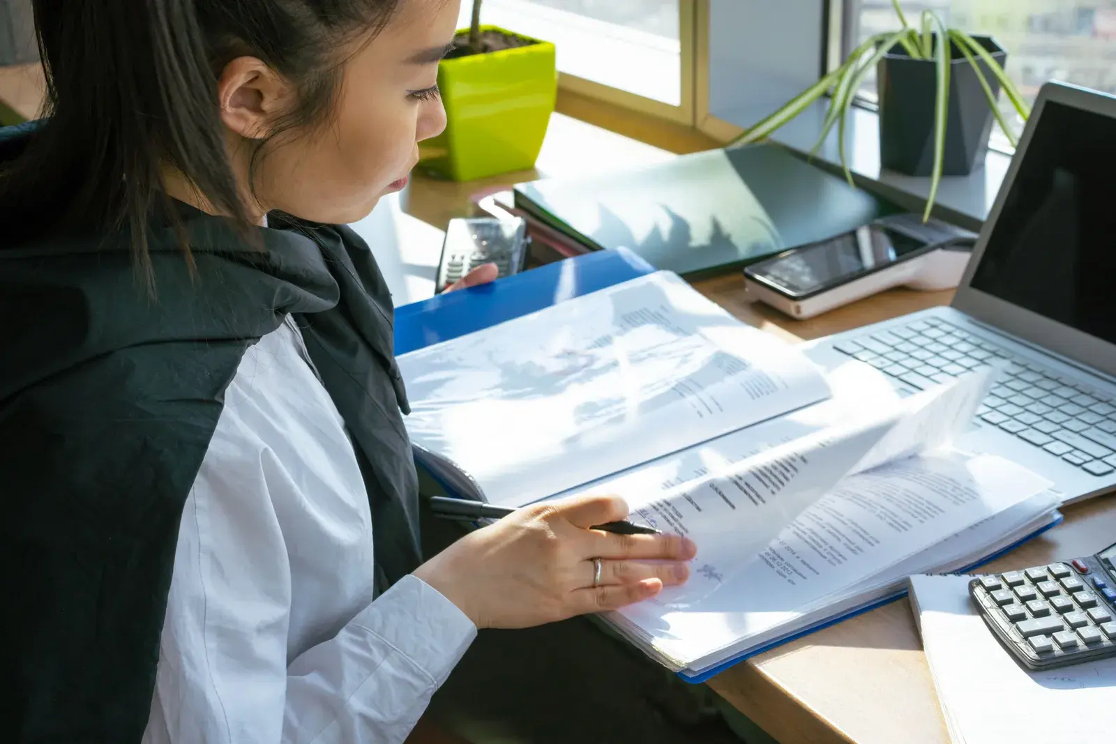 A woman reviewing a binder of printed documents with a pen at a sunlit desk beside a laptop